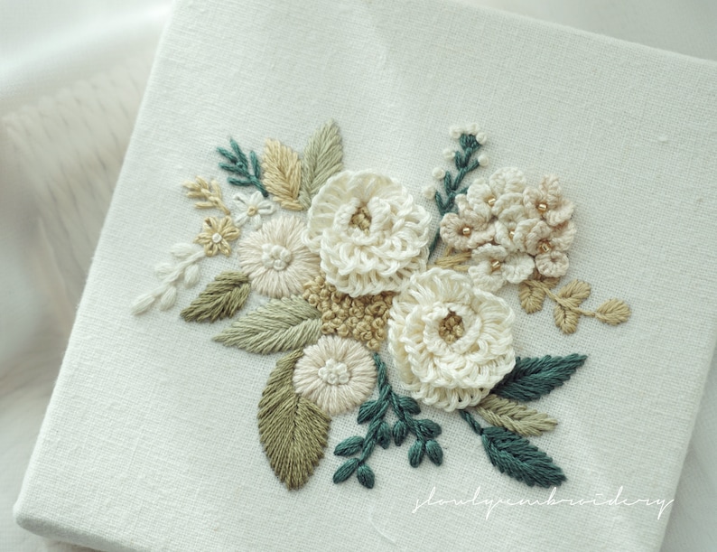 May include: A square, off-white canvas features an embroidered floral design. The embroidery includes white, cream, and tan flowers, with green and teal leaves. The design is detailed with small, gold-colored beads.