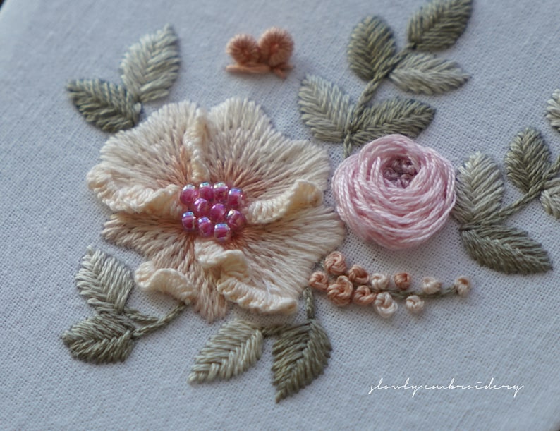 Puede incluir: Dise&ntilde;o floral bordado a mano. Se aprecia una flor grande de color melocot&oacute;n claro con detalles de cuentas rosas, una peque&ntilde;a rosa rosa y hojas verdes. Tambi&eacute;n hay una peque&ntilde;a mariposa melocot&oacute;n y otras flores peque&ntilde;as de color melocot&oacute;n sobre fondo blanco.