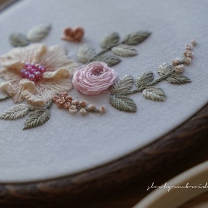 Puede incluir: Primer plano de un bordado floral en un aro de madera. El dise&ntilde;o presenta una gran flor color melocot&oacute;n con cuentas rosas, una peque&ntilde;a rosa rosa y hojas verdes, mostrando el intrincado trabajo de aguja.