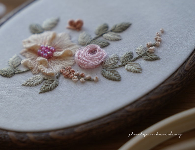 Puede incluir: Primer plano de un bordado floral en un aro de madera. El dise&ntilde;o presenta una gran flor color melocot&oacute;n con cuentas rosas, una peque&ntilde;a rosa rosa y hojas verdes, mostrando el intrincado trabajo de aguja.