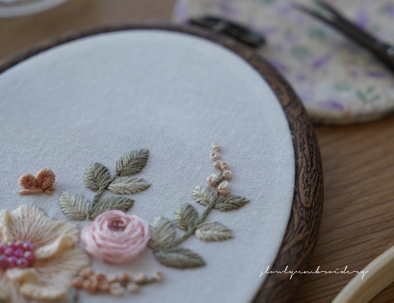 Puede incluir: Primer plano de un bastidor de bordado con un dise&ntilde;o floral. El bordado incluye una rosa rosa claro, p&eacute;talos color melocot&oacute;n y hojas verdes. El texto "slowlyembroidery" es visible. El bastidor es de madera.