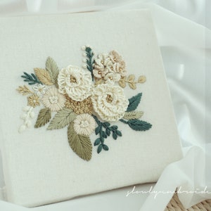 May include: A square, off-white fabric panel featuring an embroidered floral design. The embroidery includes cream-colored, beige, and green flowers and leaves. The design is centered on the panel, with the text "slowlyembroidery" in the bottom left corner.