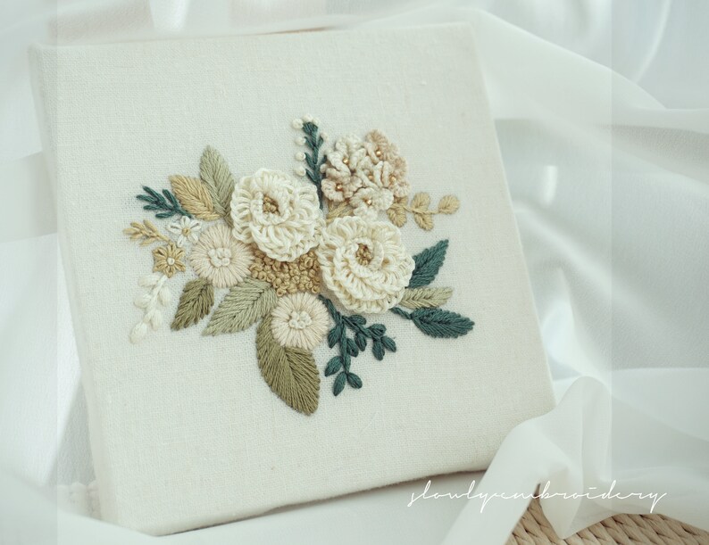 May include: A square, off-white fabric panel featuring an embroidered floral design. The embroidery includes cream-colored, beige, and green flowers and leaves. The design is centered on the panel, with the text "slowlyembroidery" in the bottom left corner.