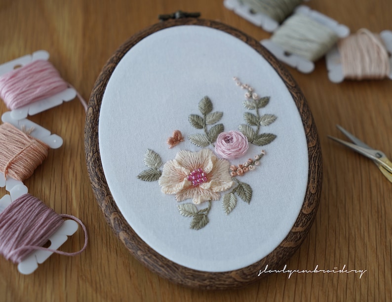 Puede incluir: Bastidor de bordado ovalado con un dise&ntilde;o floral. El bordado presenta una gran flor color melocot&oacute;n con un centro rosa, rodeada de hojas verdes y flores rosas m&aacute;s peque&ntilde;as. El bastidor tiene un acabado de vetas de madera oscura.