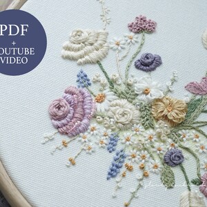 May include: An embroidered floral design in a wooden hoop. The design features a variety of flowers in shades of pink, purple, yellow, and white, with green leaves. The text "PDF + YOUTUBE VIDEO" is in a blue circle in the upper left corner.