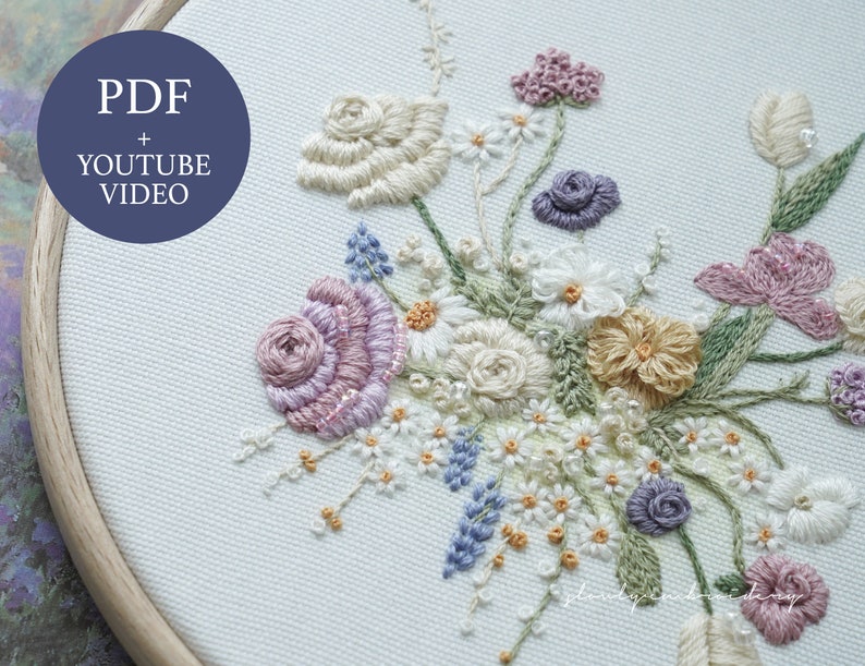 May include: An embroidered floral design in a wooden hoop. The design features a variety of flowers in shades of pink, purple, yellow, and white, with green leaves. The text "PDF + YOUTUBE VIDEO" is in a blue circle in the upper left corner.