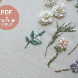 以下が含まれることがあります： さまざまな刺繍の花柄が施されたクリーム色の生地のクローズアップ。刺繍には白い花、緑の葉、その他のカラフルな花の要素が含まれています。「PDF + YOUTUBE VIDEO」というテキストは、桃色の円の中にあります。