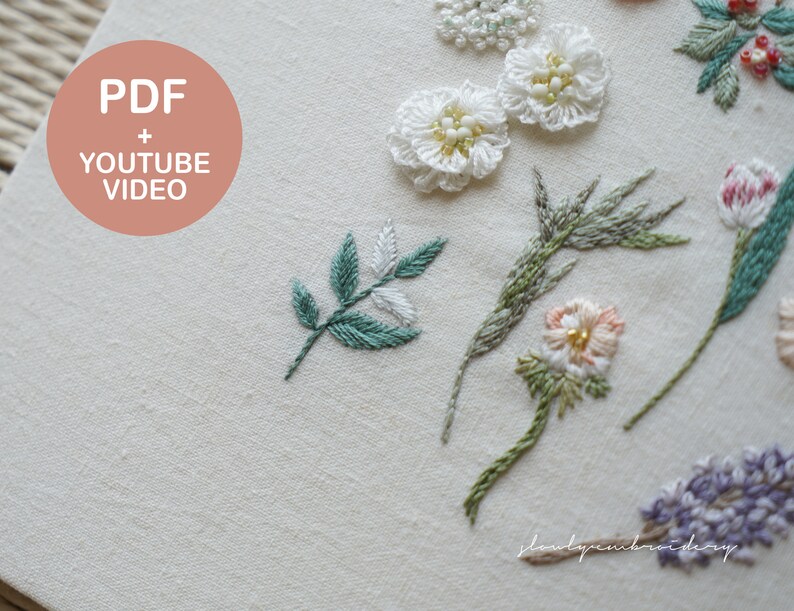 以下が含まれることがあります： さまざまな刺繍の花柄が施されたクリーム色の生地のクローズアップ。刺繍には白い花、緑の葉、その他のカラフルな花の要素が含まれています。「PDF + YOUTUBE VIDEO」というテキストは、桃色の円の中にあります。