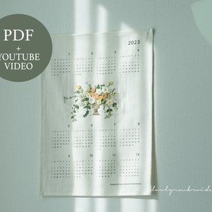 以下が含まれることがあります： 壁に掛けられたクリーム色の2023年カレンダー。花の刺繍が施されています。カレンダーには、黄色、桃色、白色の花で飾られた装飾的な花瓶があります。「PDF + YOUTUBE VIDEO」というテキストが左上に円で表示されています。