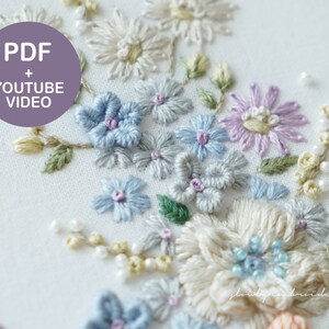 May include: Close-up of an embroidered floral design featuring various flowers in shades of blue, purple, and cream, with green leaves and pearl-like beads. The text "PDF + YOUTUBE VIDEO" is in a purple circle.