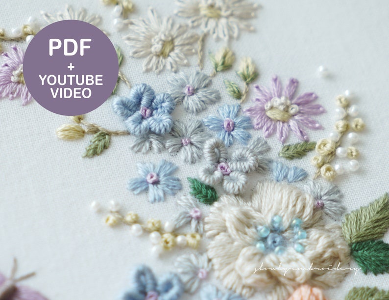 May include: Close-up of an embroidered floral design featuring various flowers in shades of blue, purple, and cream, with green leaves and pearl-like beads. The text "PDF + YOUTUBE VIDEO" is in a purple circle.