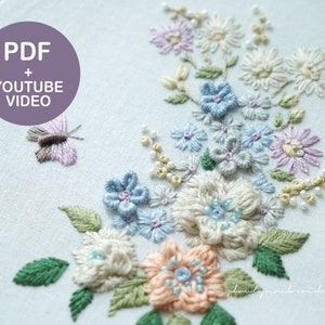 May include: Embroidered floral design in a wooden hoop. The design features pastel-colored flowers, including blue, peach, and lavender, with green leaves and a small butterfly. The text "PDF + YOUTUBE VIDEO" is in a purple circle.