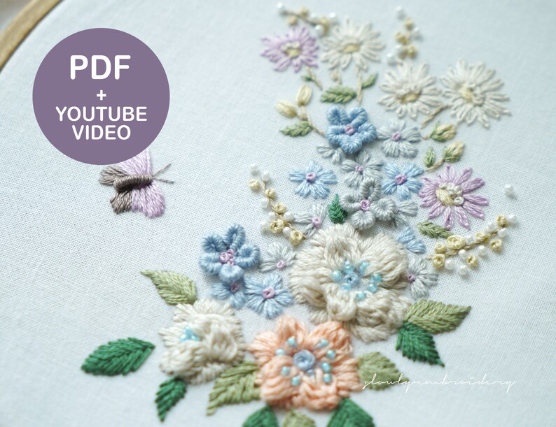 May include: Embroidered floral design in a wooden hoop. The design features pastel-colored flowers, including blue, peach, and lavender, with green leaves and a small butterfly. The text "PDF + YOUTUBE VIDEO" is in a purple circle.