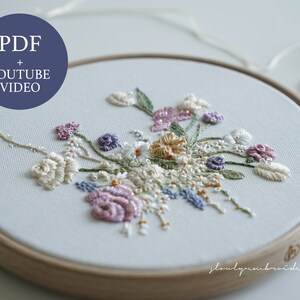 May include: An embroidery hoop with a floral design. The design features a variety of flowers in shades of white, pink, purple, and yellow, with green leaves. The text "PDF + YOUTUBE VIDEO" is in a blue circle.