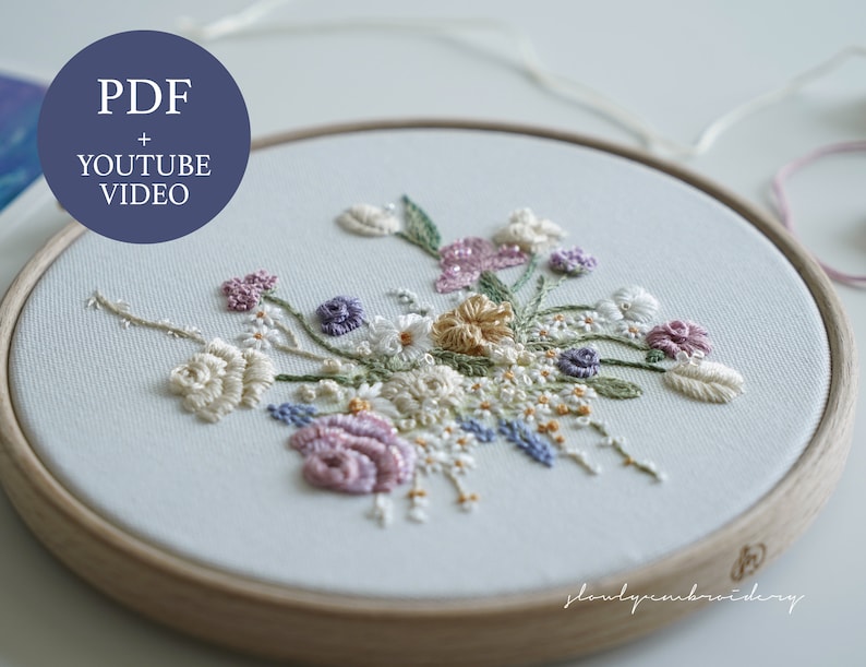 May include: An embroidery hoop with a floral design. The design features a variety of flowers in shades of white, pink, purple, and yellow, with green leaves. The text "PDF + YOUTUBE VIDEO" is in a blue circle.