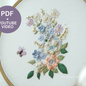 May include: An embroidery hoop featuring a floral design with a butterfly. The design includes flowers in shades of blue, peach, and cream, with green leaves. The text "PDF + YOUTUBE VIDEO" is in a purple circle.