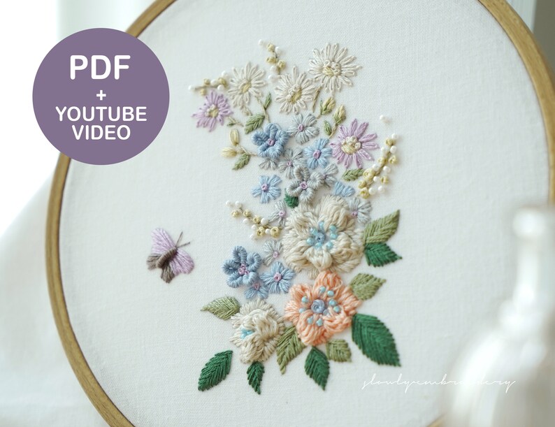 May include: An embroidery hoop featuring a floral design with a butterfly. The design includes flowers in shades of blue, peach, and cream, with green leaves. The text "PDF + YOUTUBE VIDEO" is in a purple circle.