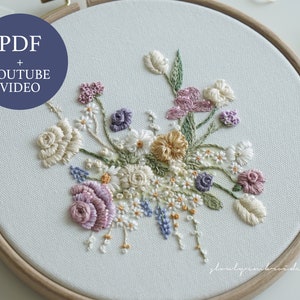 May include: An embroidered floral design in a wooden hoop. The artwork features a bouquet of various flowers in shades of cream, purple, yellow, and blue. The text "PDF + YOUTUBE VIDEO" is in a blue circle in the upper left corner.