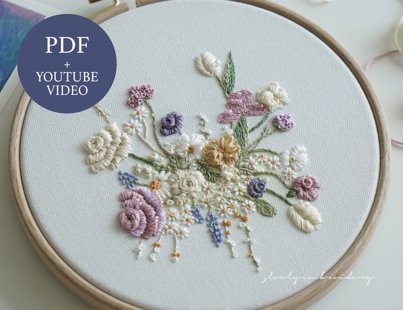 May include: An embroidered floral design in a wooden hoop. The artwork features a bouquet of various flowers in shades of cream, purple, yellow, and blue. The text "PDF + YOUTUBE VIDEO" is in a blue circle in the upper left corner.
