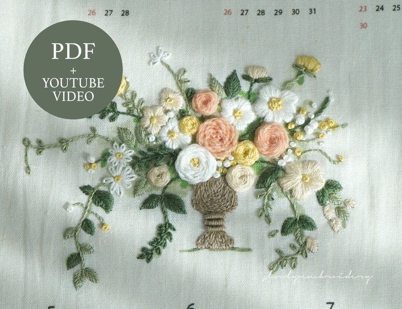 以下が含まれることがあります： 白い、桃色、黄色の花束が茶色の花瓶に入った刺繍デザイン。緑の葉とつるが花を囲んでいます。「PDF + YOUTUBE VIDEO」というテキストが緑色の円の中にあります。