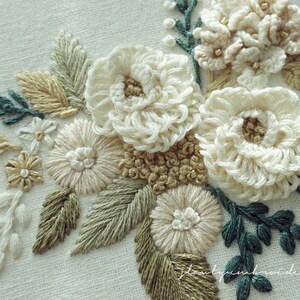 May include: Close-up of an embroidered floral design featuring cream-colored flowers with tan centers and accents. The embroidery includes green and tan leaves and stems, creating a natural, textured appearance. The background is a light, off-white fabric.