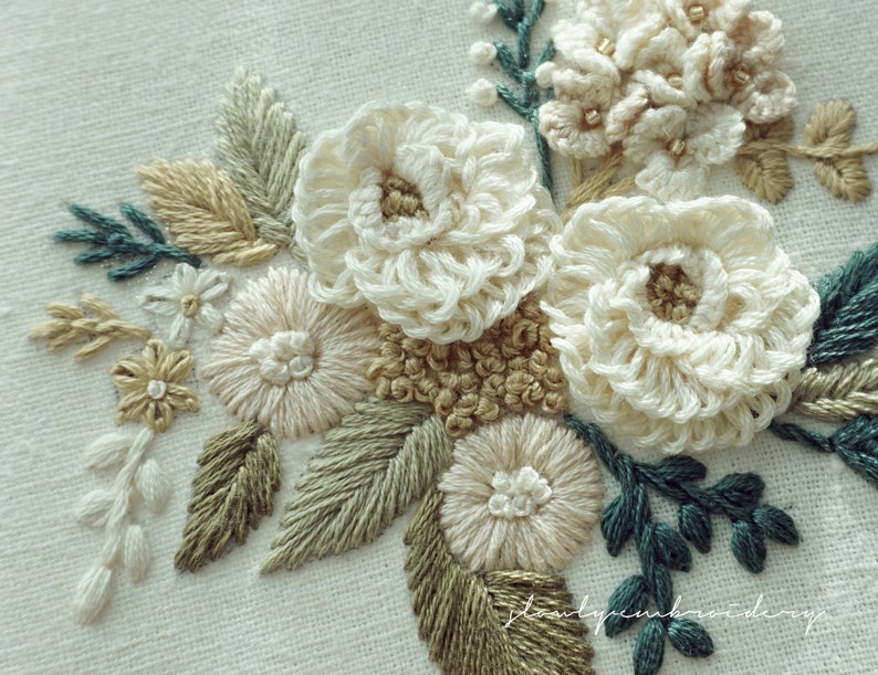 May include: Close-up of an embroidered floral design featuring cream-colored flowers with tan centers and accents. The embroidery includes green and tan leaves and stems, creating a natural, textured appearance. The background is a light, off-white fabric.