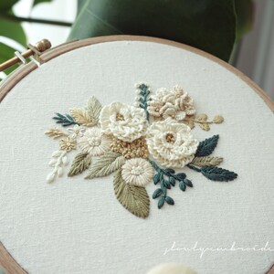 May include: A floral embroidery hoop featuring a cream-colored flower bouquet with green and tan leaves. The embroidery is set against a white fabric background, framed by a wooden hoop.