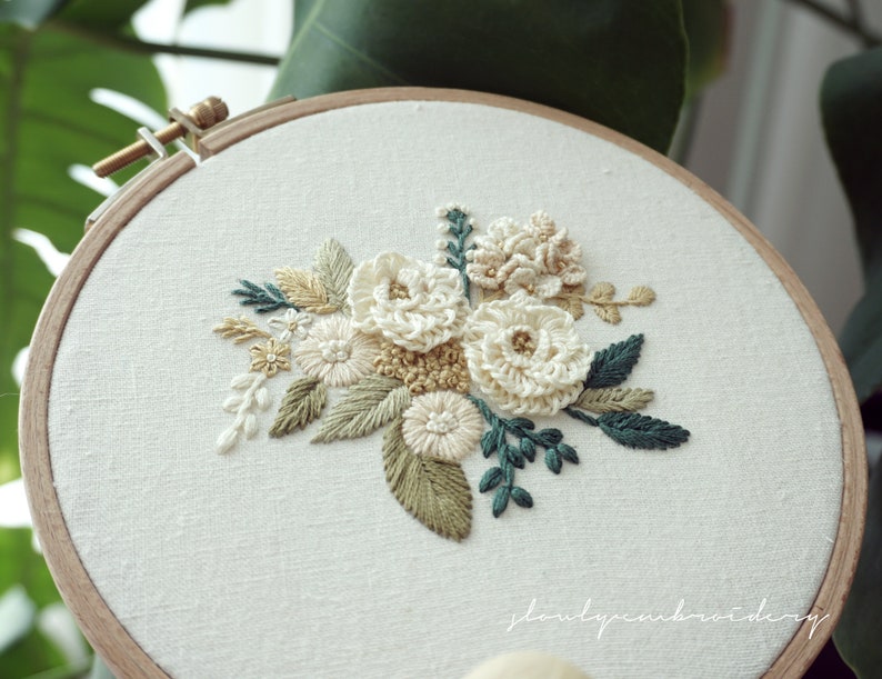 May include: A floral embroidery hoop featuring a cream-colored flower bouquet with green and tan leaves. The embroidery is set against a white fabric background, framed by a wooden hoop.
