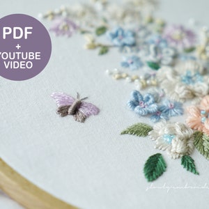 May include: Close-up of an embroidery hoop with a floral design and a butterfly. The design features pastel-colored flowers in shades of pink, blue, and white, with green leaves. A small butterfly is stitched near the flowers. Text on the image reads "PDF + YOUTUBE VIDEO".