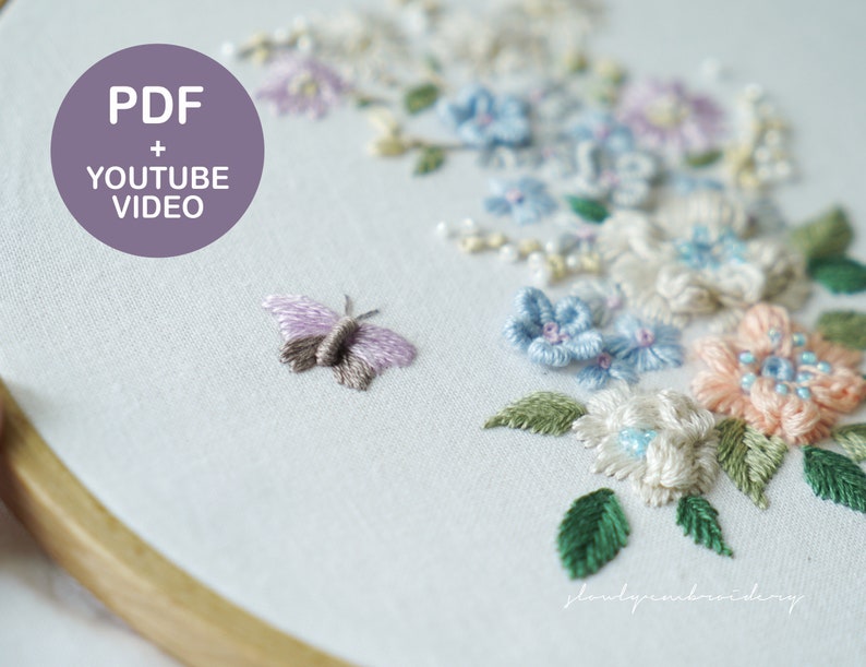 May include: Close-up of an embroidery hoop with a floral design and a butterfly. The design features pastel-colored flowers in shades of pink, blue, and white, with green leaves. A small butterfly is stitched near the flowers. Text on the image reads "PDF + YOUTUBE VIDEO".