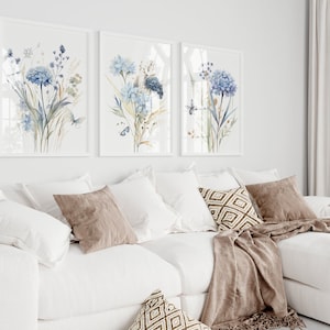 May include: Three framed watercolor paintings of blue flowers with green stems and leaves. The paintings are hung on a white wall above a white couch with brown throw pillows and a brown blanket.