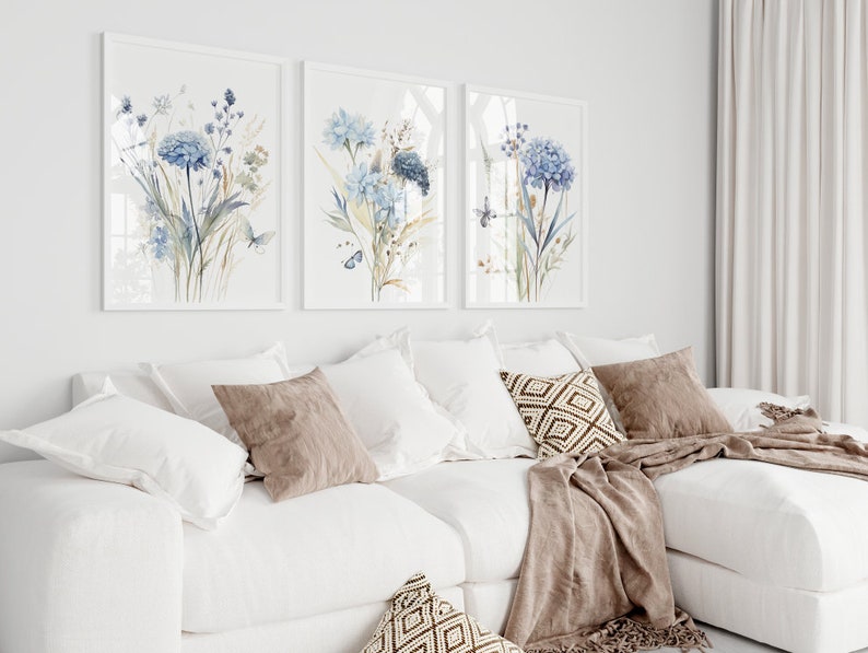 May include: Three framed watercolor paintings of blue flowers with green stems and leaves. The paintings are hung on a white wall above a white couch with brown throw pillows and a brown blanket.