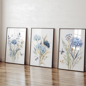 May include: Three framed watercolor prints featuring blue flowers and butterflies. The prints are in black frames and are arranged in a row on a wooden floor.