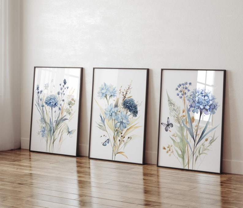 May include: Three framed watercolor prints featuring blue flowers and butterflies. The prints are in black frames and are arranged in a row on a wooden floor.
