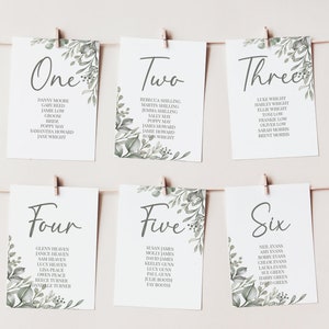 May include: Six white cards with green foliage designs and handwritten numbers one to six. Each card lists names for a wedding reception seating chart.