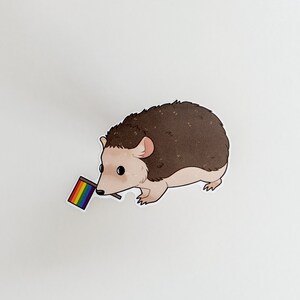 May include: A brown and white hedgehog holding a small rainbow flag in its mouth.
