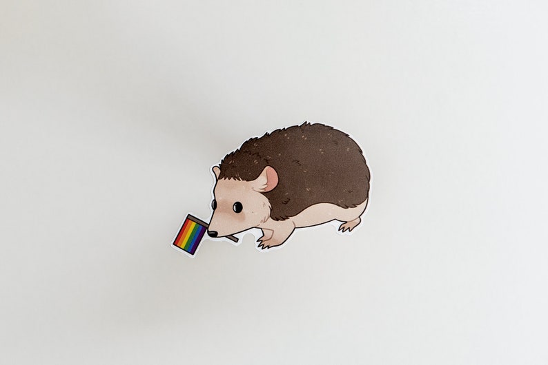 May include: A brown and white hedgehog holding a small rainbow flag in its mouth.