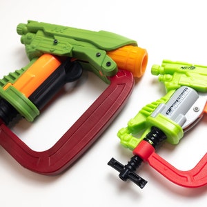May include: Two toy guns, one green and orange, the other green and silver, are attached to red C-clamps. The green and orange gun has the word "NERF" on the side. The green and silver gun has the words "SUPER SOAKER" on the side. The red C-clamps have the word "CLAMPDOR" on the side.