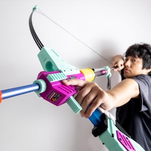 May include: A colorful toy bow and arrow with a blue, pink, and green color scheme. The bow is made of plastic and has a string attached to it. The arrow is also made of plastic and has a black tip.