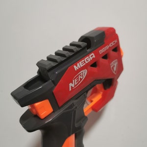 May include: A black and red Nerf Mega Bigshock toy blaster with an orange trigger and a black rail on top. The blaster has a red and black grip and the words "MEGA BIGSHOCK" are printed on the side.