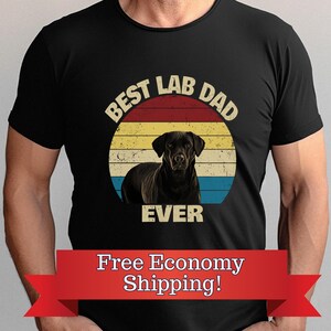 May include: Black t-shirt with a graphic of a black Labrador Retriever inside a circular design with a red, yellow, and blue striped background. The text "BEST LAB DAD EVER" is printed above and below the dog image.