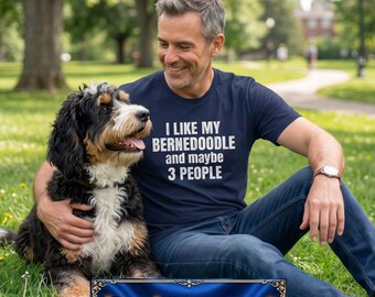 May include: A navy blue t-shirt with the text "I like my Bernedoodle and maybe 3 people" in white. The shirt is worn by a person sitting on the grass with a Bernedoodle dog. The dog has black, brown, and white fur.