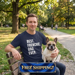 May include: A man wearing a navy blue t-shirt with the text "BEST. FRENCHIE. DAD. EVER." is seated on a park bench, holding a French Bulldog. The dog has a white and brown coat. The background shows a park with trees and a walking path.