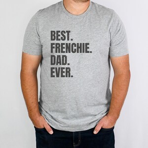 May include: A gray t-shirt with the text "BEST. FRENCHIE. DAD. EVER." in black. The shirt is short-sleeved and worn by a person. The person is wearing dark blue jeans.