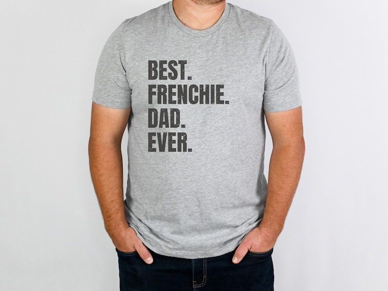 May include: A gray t-shirt with the text "BEST. FRENCHIE. DAD. EVER." in black. The shirt is short-sleeved and worn by a person. The person is wearing dark blue jeans.