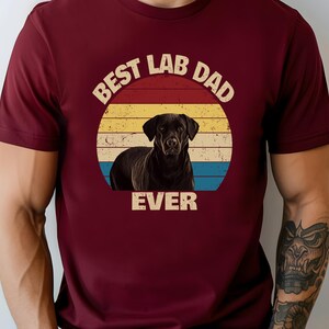 May include: A maroon t-shirt with a graphic of a black Labrador Retriever set against a retro sunset design. The text reads "BEST LAB DAD EVER". The shirt is a solid colour.