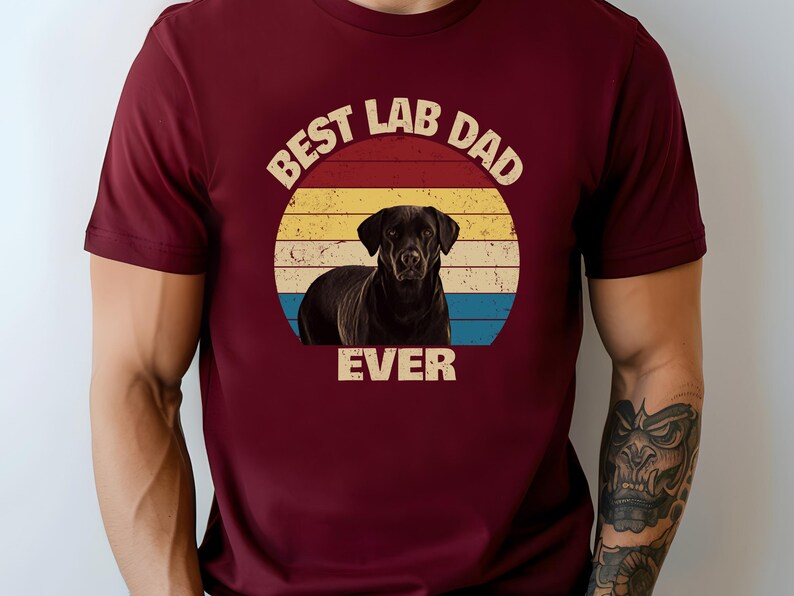 May include: A maroon t-shirt with a graphic of a black Labrador Retriever set against a retro sunset design. The text reads "BEST LAB DAD EVER". The shirt is a solid colour.