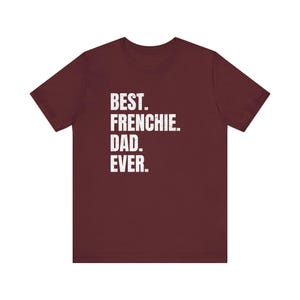 May include: A maroon t-shirt with the text "BEST. FRENCHIE. DAD. EVER." in white block letters. The shirt is a crew neck style and appears to be made of a soft material. The text is centered on the front of the shirt.