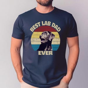 以下が含まれることがあります： ネイビーブルーのTシャツ。レトロなデザインで、ラブラドールレトリバーと「BEST LAB DAD EVER」の文字が赤、黄、青のストライプの円の中に描かれています。クラシックなクルーネックのシャツです。