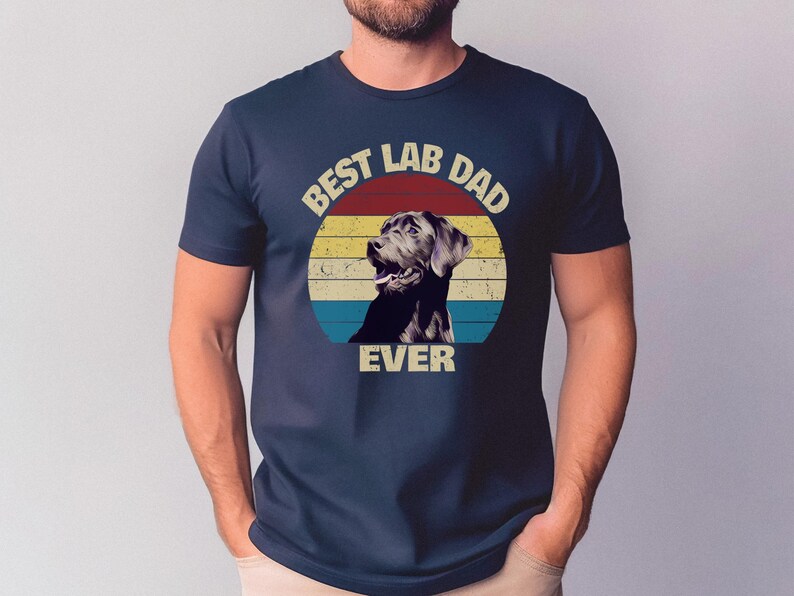 以下が含まれることがあります： ネイビーブルーのTシャツ。レトロなデザインで、ラブラドールレトリバーと「BEST LAB DAD EVER」の文字が赤、黄、青のストライプの円の中に描かれています。クラシックなクルーネックのシャツです。